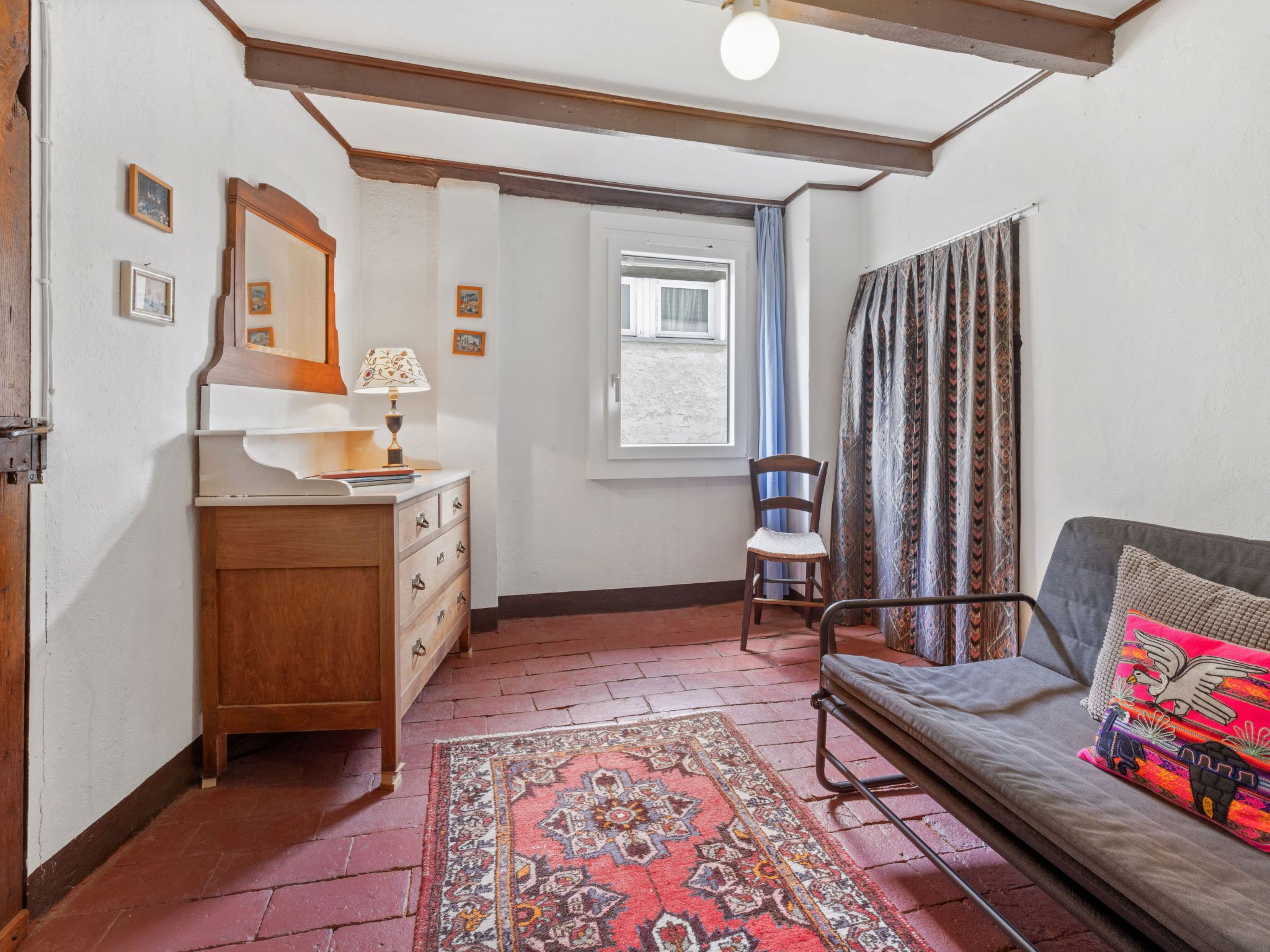 A cozy room with a wooden dresser, a chair, a sofa with colorful cushions, a patterned rug, and a window with curtains.