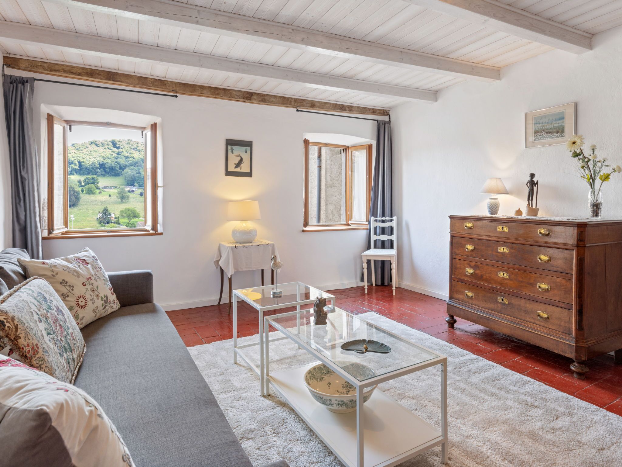 A cozy living room with a sofa, a glass coffee table, a wooden dresser, and two windows offering a view of greenery.