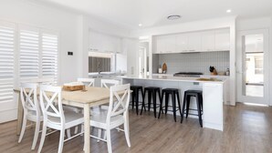 A modern kitchen with white cabinetry, a central island with stools, and a dining area with white chairs. - Newly Built Luxury Modern Home (Dunsborough)