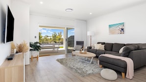 A modern living room with a grey sofa, a coffee table, and a TV on the wall. There is a balcony with a view of trees and a railing. - Newly Built Luxury Modern Home (Dunsborough)