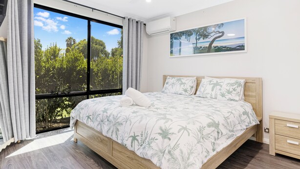 Een slaapkamer met een groot raam, een bed met een bloemenbedekking, een nachtkastje en een luchtverkoeler aan de muur.