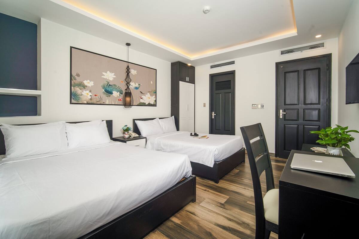 Quarto de hotel moderno com duas camas, uma mesa com um laptop, uma grande pintura floral e uma porta escura.