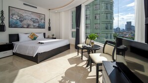 Quarto de hotel moderno com uma cama grande, uma mesa redonda pequena e vista para a cidade através de amplas janelas.
