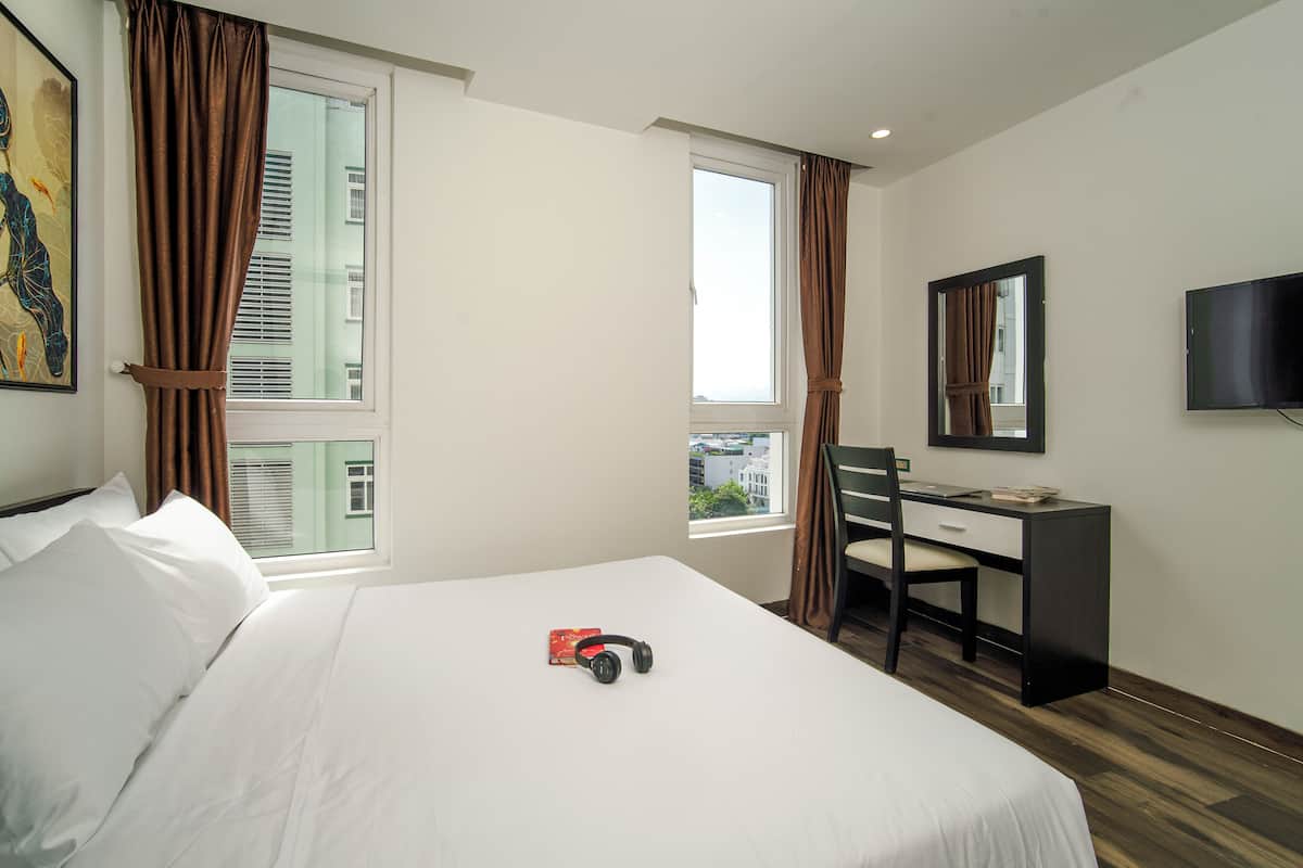 Quarto de hotel com cama, escrivaninha, cadeira, televisão e duas janelas com cortinas.