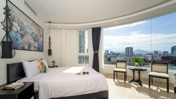 Quarto de hotel moderno com uma cama grande, uma mesa pequena, duas cadeiras, uma janela ampla com vista para a cidade e um quadro na parede.