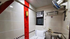 A small, clean room with a bed, a red accent wall, a window with an air conditioner, a bedside table with a phone, and a red toilet seat. - NEW PEKING GUEST HOUSE (Kowloon)