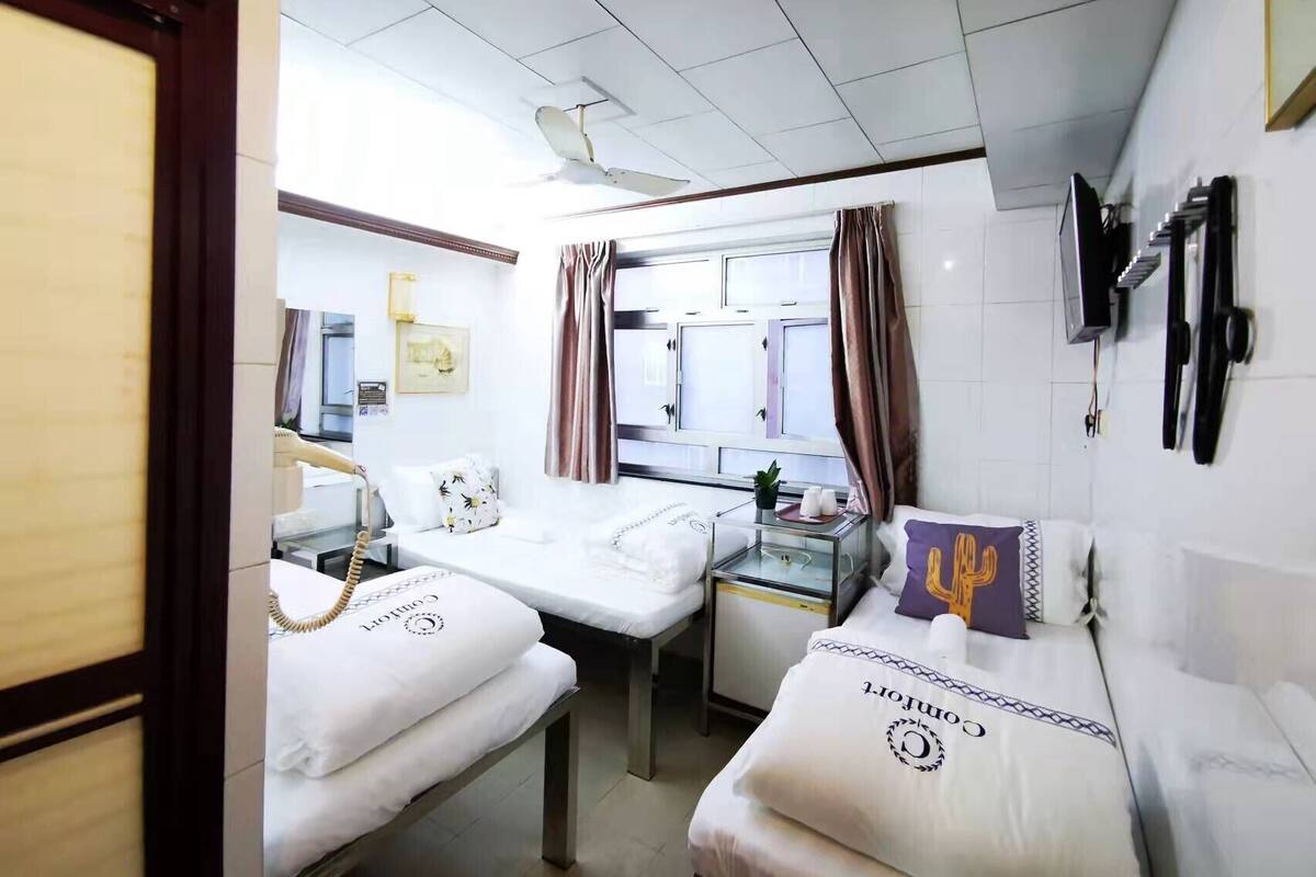Ein Hotelzimmer mit zwei Betten, einem Schreibtisch, einem Deckenventilator, einem Fenster mit Vorhängen und einem an der Wand befestigten Fernseher.