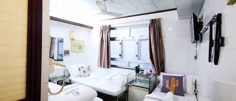 A hotel room with two beds, a desk, a ceiling fan, a window with curtains, and a TV mounted on the wall.