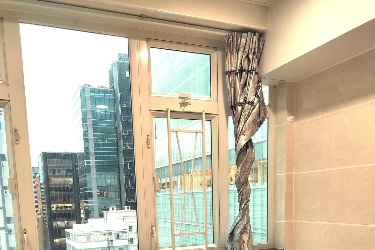 Ein Hotelzimmer mit Bett, einem Fenster mit Vorhängen, Blick auf Stadtgebäude und einer gefliesten Wand.