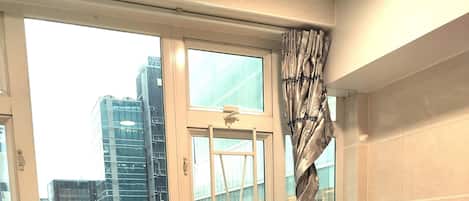 A hotel room with a bed, a window with curtains, a view of city buildings, and a tiled wall.