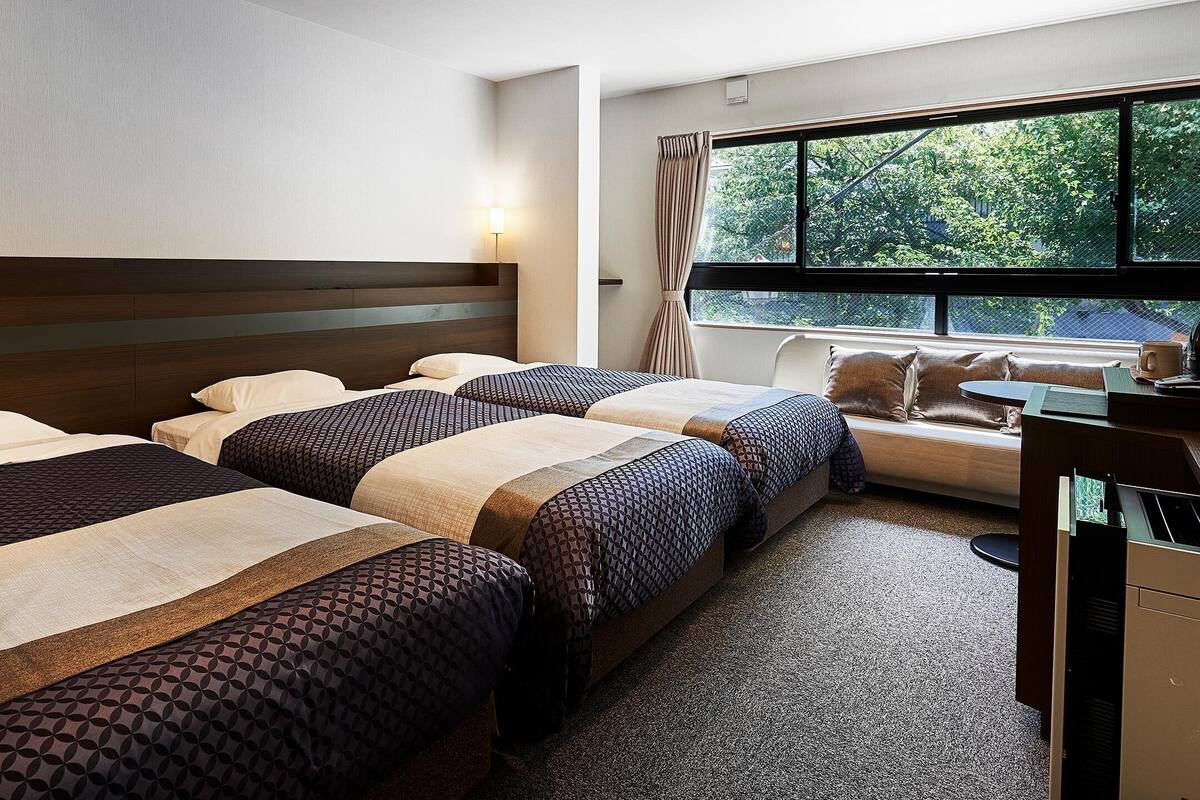 A hotel room with two beds, a desk, and a window with a view of trees.