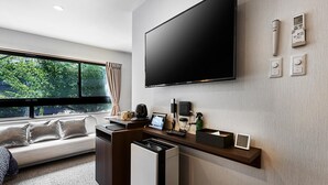 A modern hotel room with a large flat-screen TV mounted on the wall, a desk with a coffee maker, a small clock, and a window with curtains.