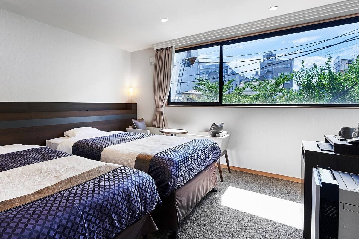 A hotel room with two beds, a desk, a chair, a window with a view of buildings, and a ceiling fan.