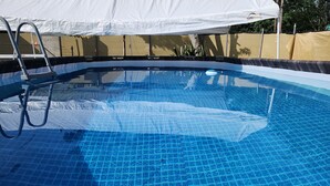 Une piscine à eau claire dont le fond est recouvert de carreaux bleus, avec une échelle et un auvent blanc au-dessus.