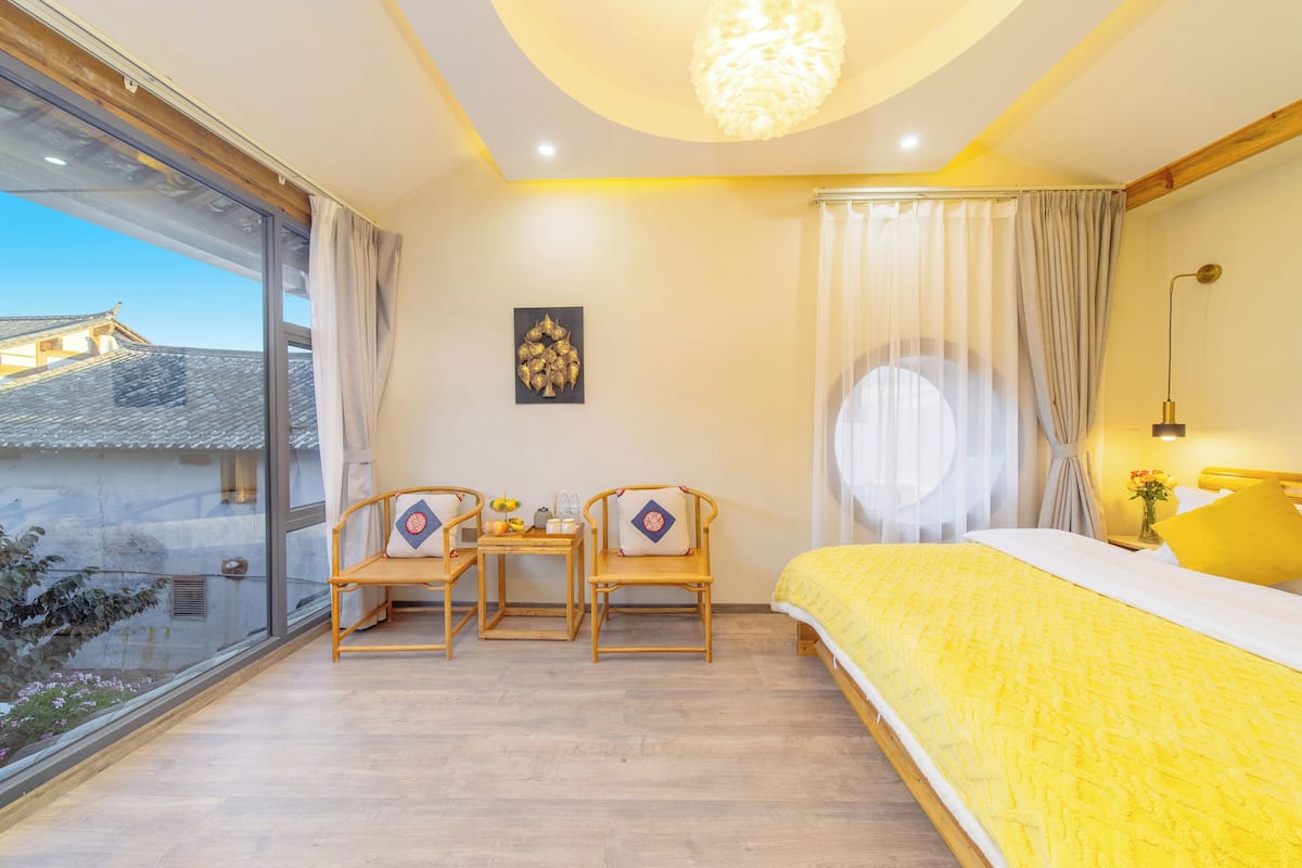 A bedroom with a large window, a bed with a yellow bedspread, two wooden chairs, a small table with a vase, and a wall-mounted lamp.