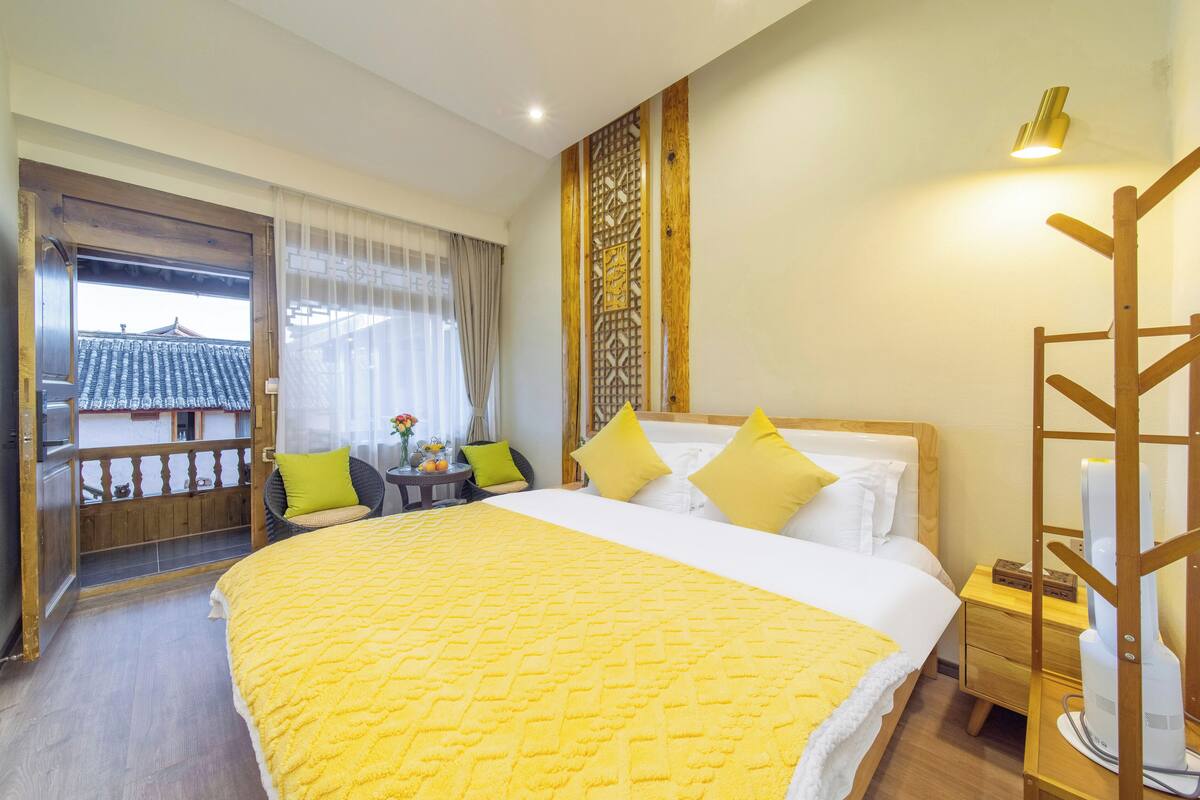 A hotel room with a large bed, a wooden nightstand, a chair with cushions, a view of a traditional building through a wooden door, and a decorative wall panel.