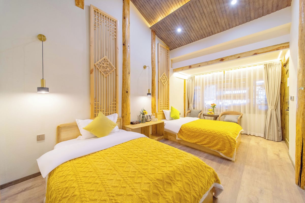 A hotel room with two beds, wooden beams, a ceiling light, a window with curtains, and a small table with a vase of flowers.