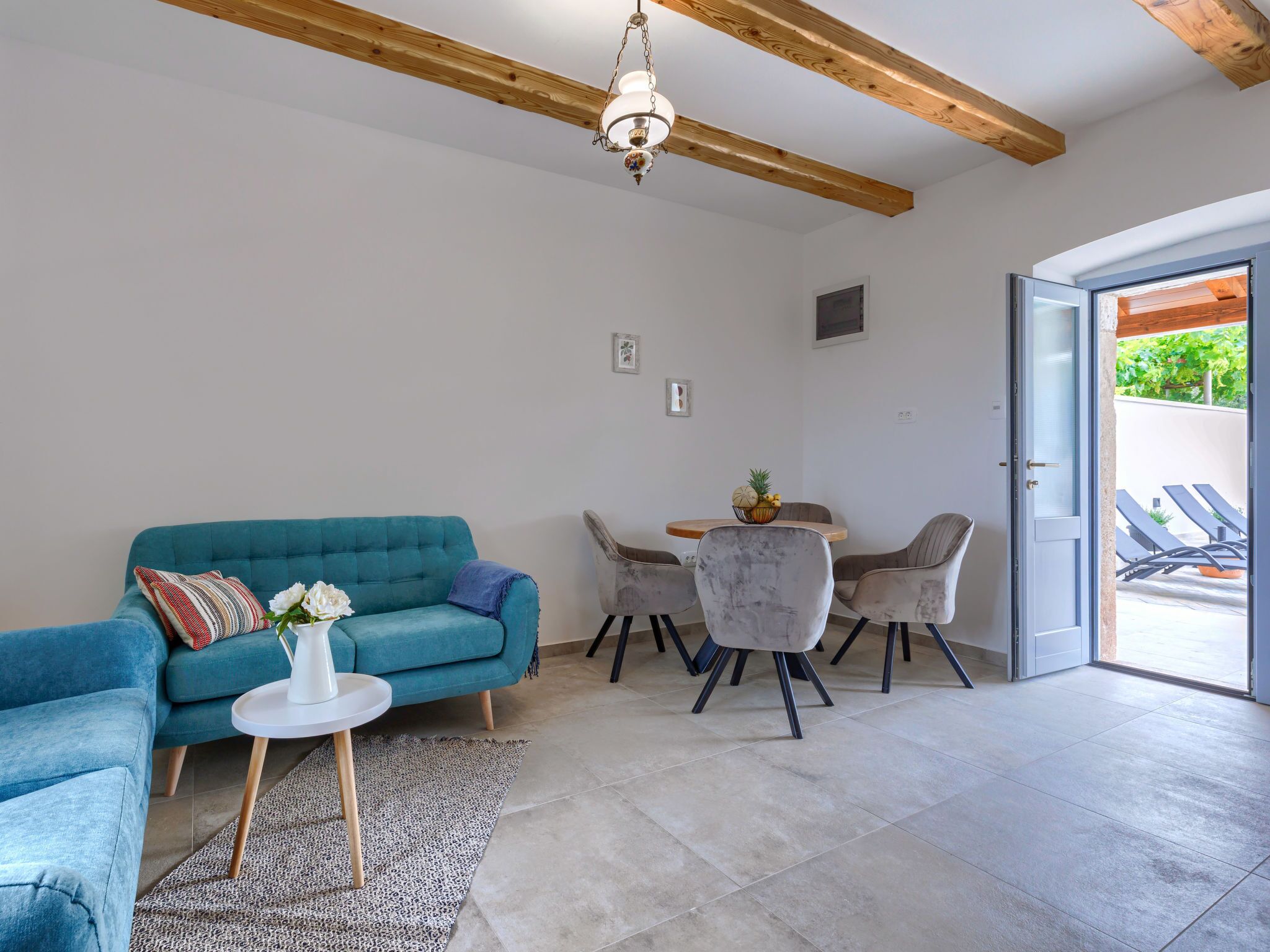Ein modernes Wohnzimmer mit einem türkisfarbenen Sofa, einem kleinen runden Tisch mit Stühlen und einer Tür zu einem Außenbereich mit Liegestühlen.