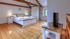 A bedroom with a wooden ceiling, a bed with white linens, a nightstand, a desk with a television, and a stone wall with a window. - Villa Lucky by Interhome (Poreč)