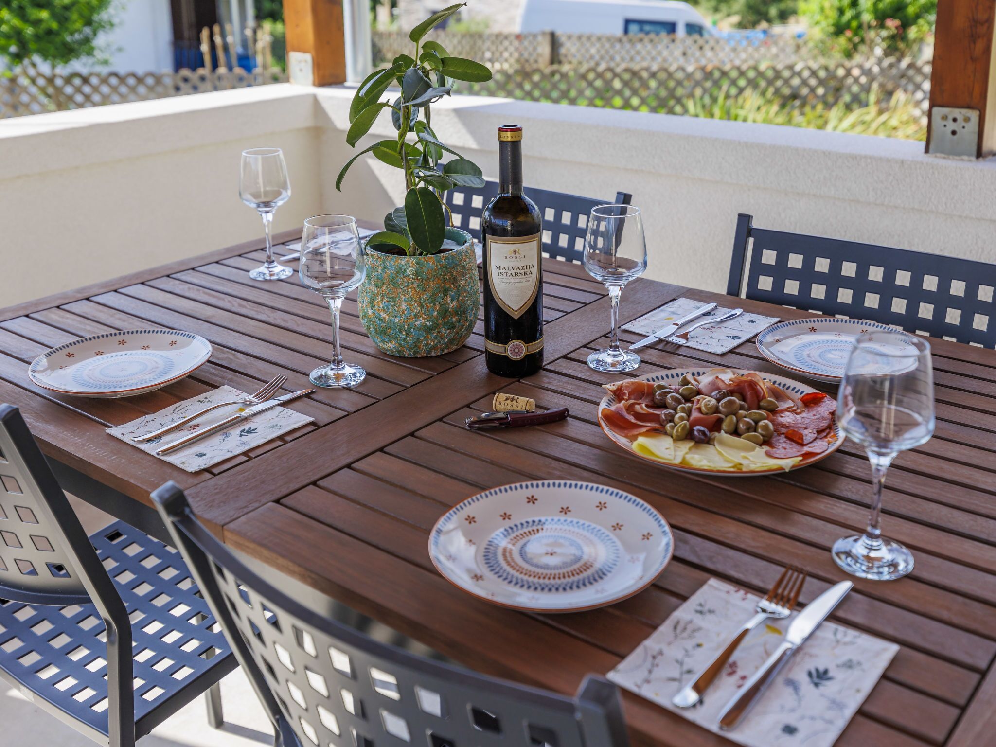 A wooden table set for a meal with plates, cutlery, a bottle of wine, and a potted plant.