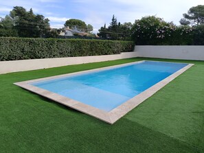 A rectangular swimming pool with a light blue interior, surrounded by artificial green grass and bordered by a light-colored stone edge.
