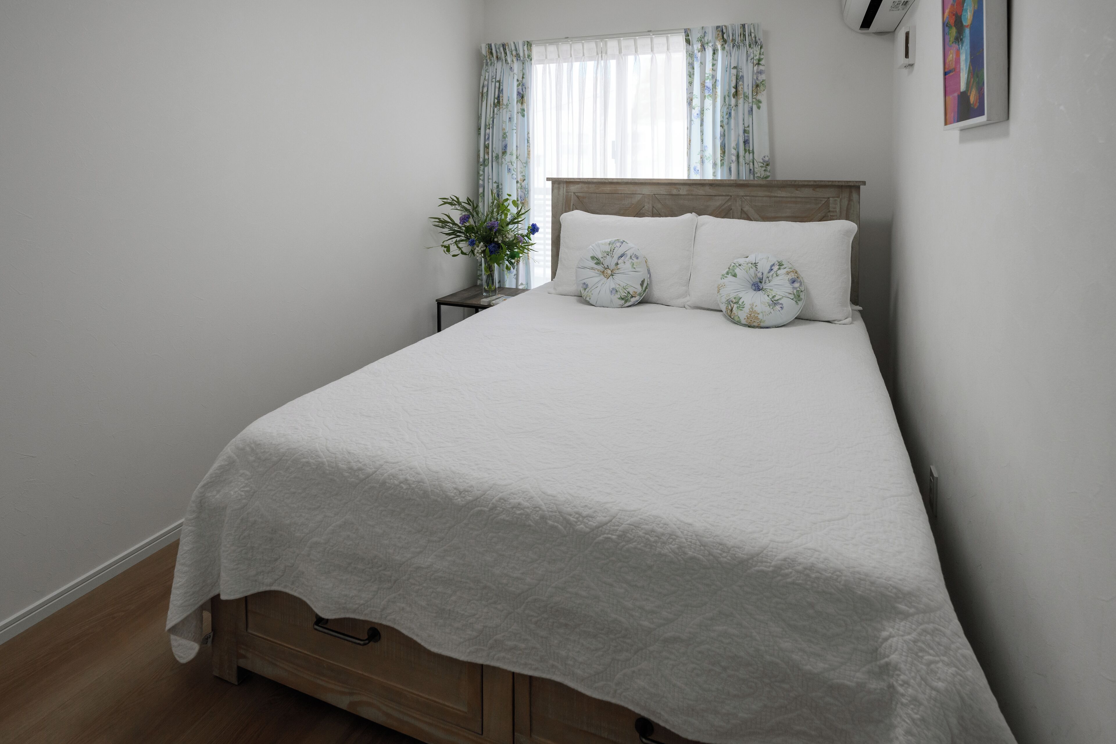 Un lit bien fait, recouvert d’une couvre-lit blanche et agrémenté d’oreillers à motifs floraux, une table de chevet avec un vase de fleurs et une fenêtre ornée de rideaux légers.