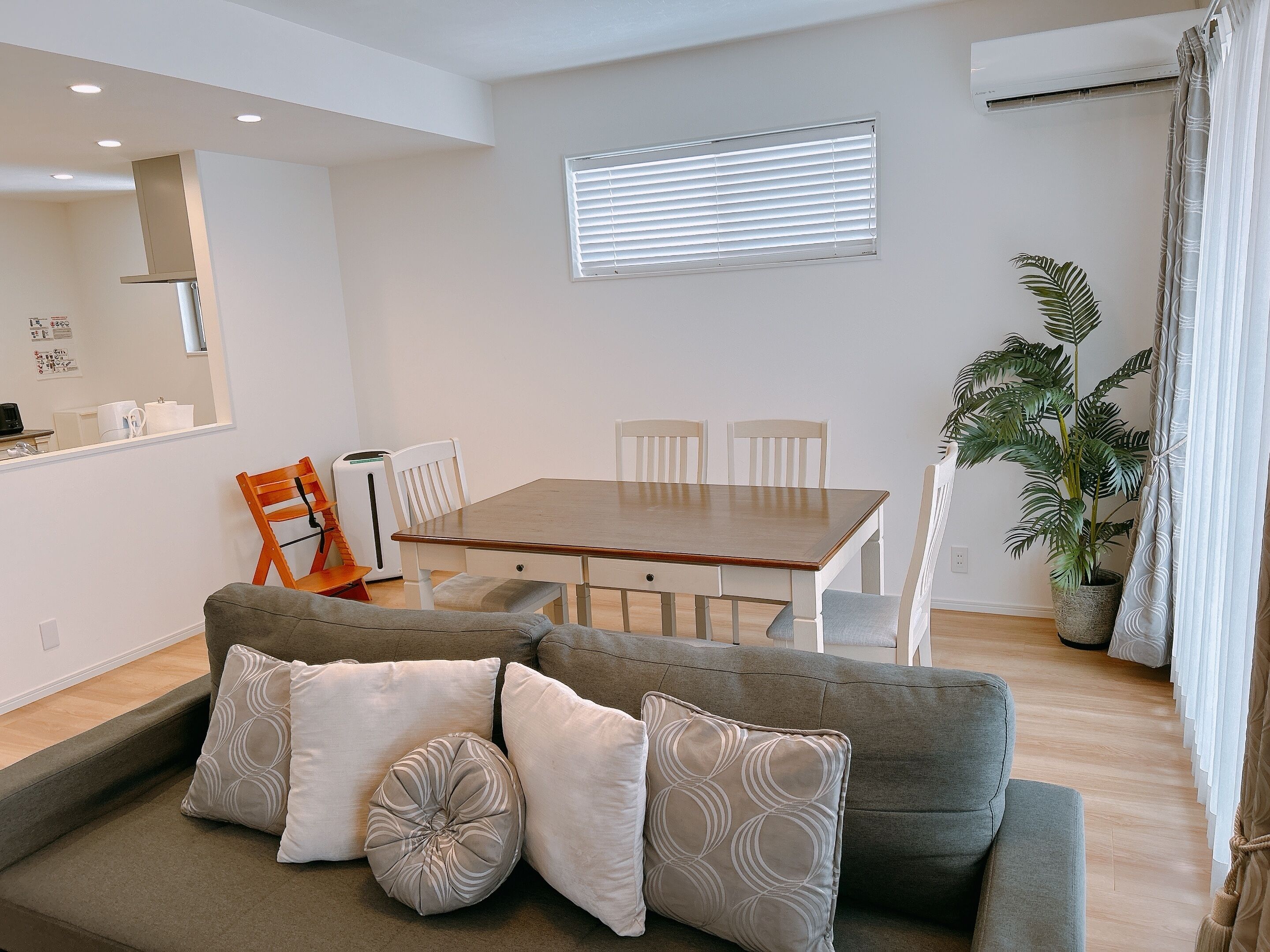 A modern living room with a wooden dining table, white chairs, a grey sofa with patterned cushions, a potted plant, and a large mirror on the wall.