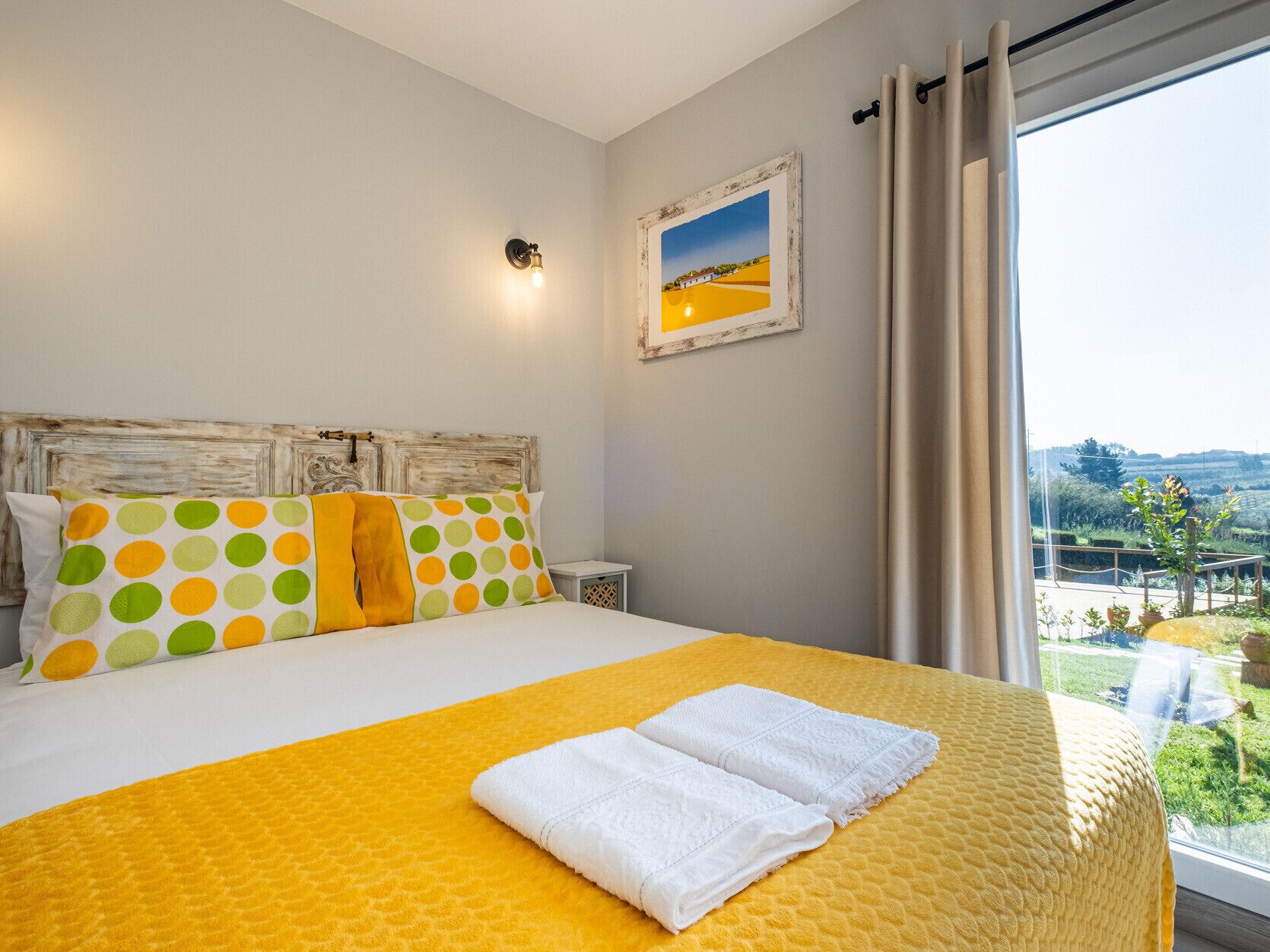 A bedroom with a bed, pillows, a nightstand, a wall-mounted light, and a window with curtains.