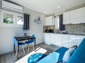 Een compacte keuken met witte kastjes, een magnetron en een decoratieve spatwand. Er is een eethoek met een donkere tafel en blauwe stoelen, en een blauwe bank in de woonkamer.