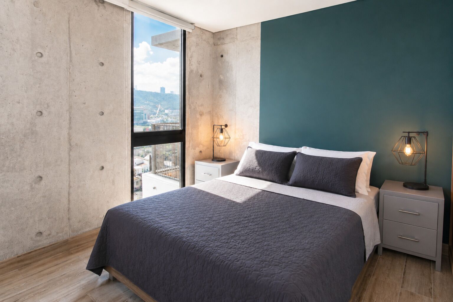 A bedroom with a large bed, two bedside tables, a view through a window, and a teal accent wall.