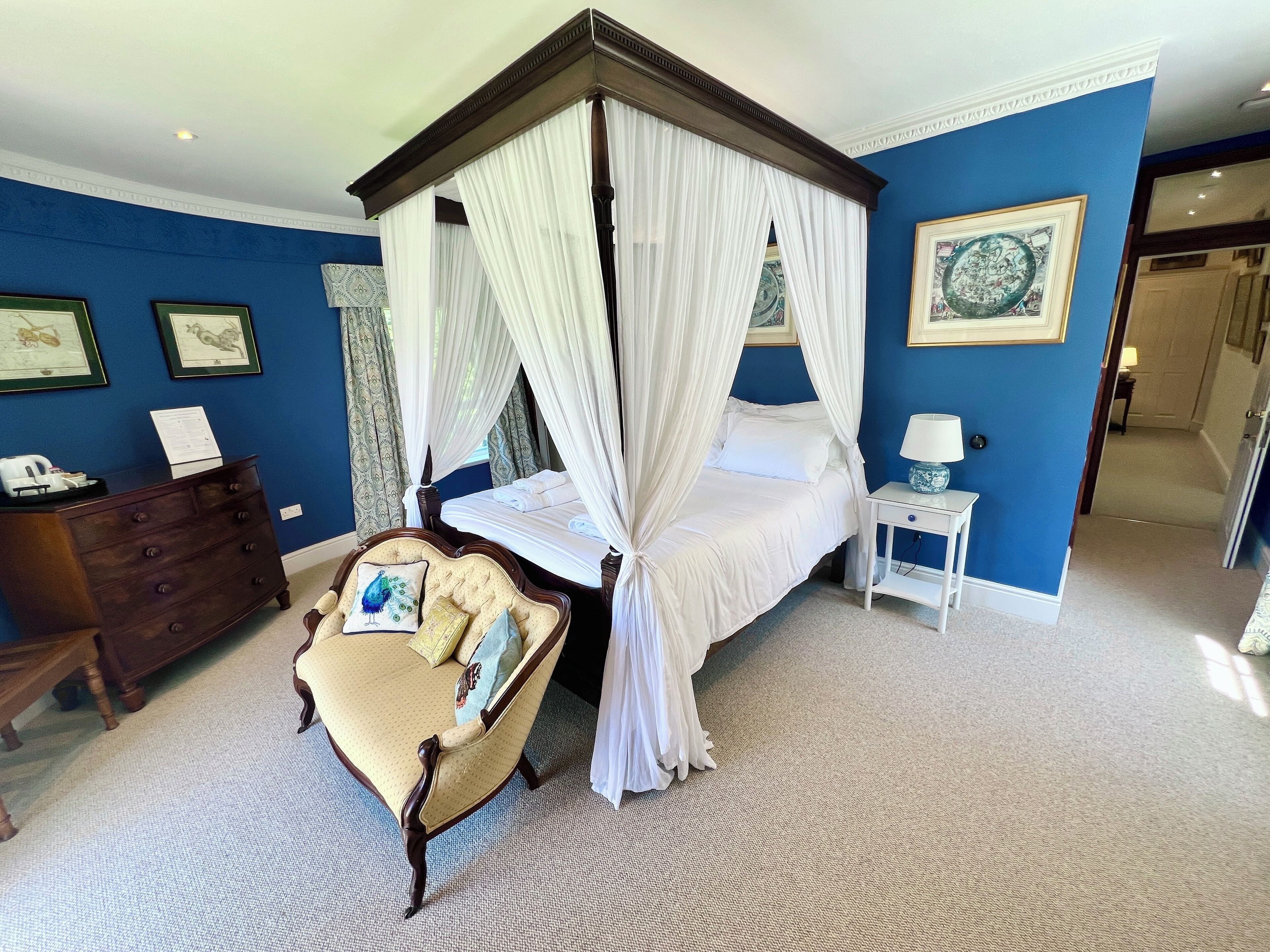 A four-poster canopy bed with white linens, a tufted armchair with patterned cushions, a nightstand with a lamp, and framed artwork on the walls.