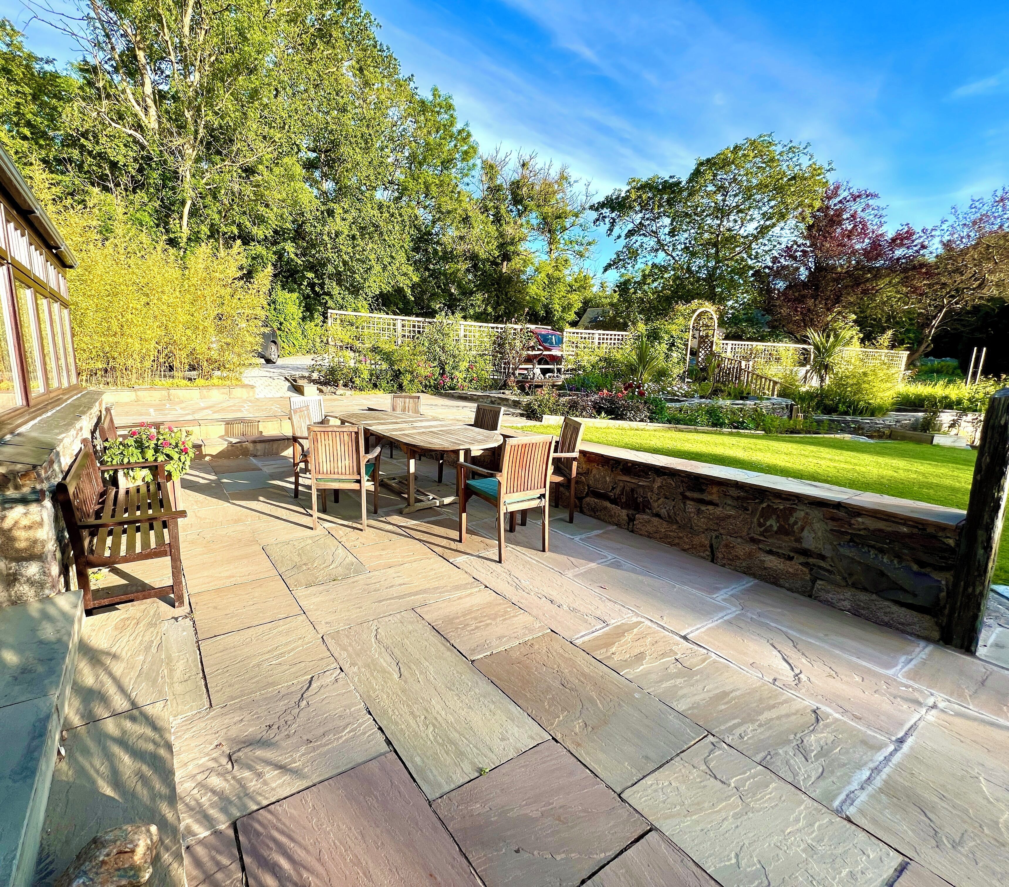 A patio area with stone flooring, wooden furniture, and a view of a garden with a stone wall and trees.