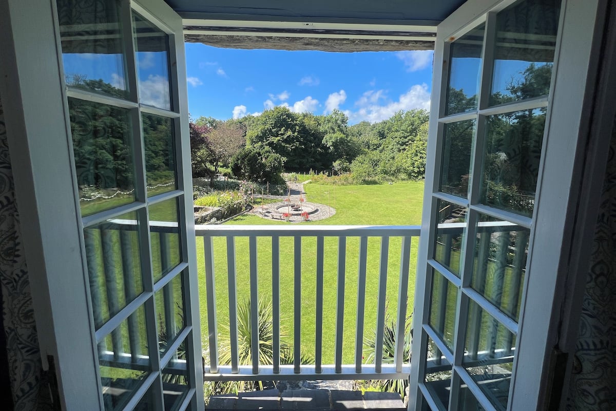 A view from a room with a balcony, featuring a well-maintained lawn, a circular flower bed, and a variety of trees and shrubs.
