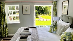 A hotel room with a bed, a window with curtains, a framed picture, and a view of a garden. - The Mill House (Ballasalla)