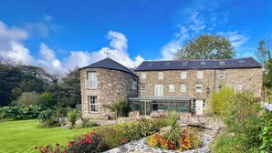 A stone building with a circular tower, multiple windows, and a garden with various plants and flowers. - The Mill House (Ballasalla)
