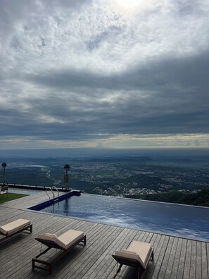 An infinity pool with lounge chairs, overlooking a cityscape and mountains under a cloudy sky.