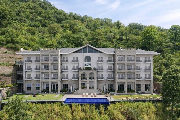 A multi-story hotel with a central courtyard, a swimming pool, and a lush green hillside backdrop.
