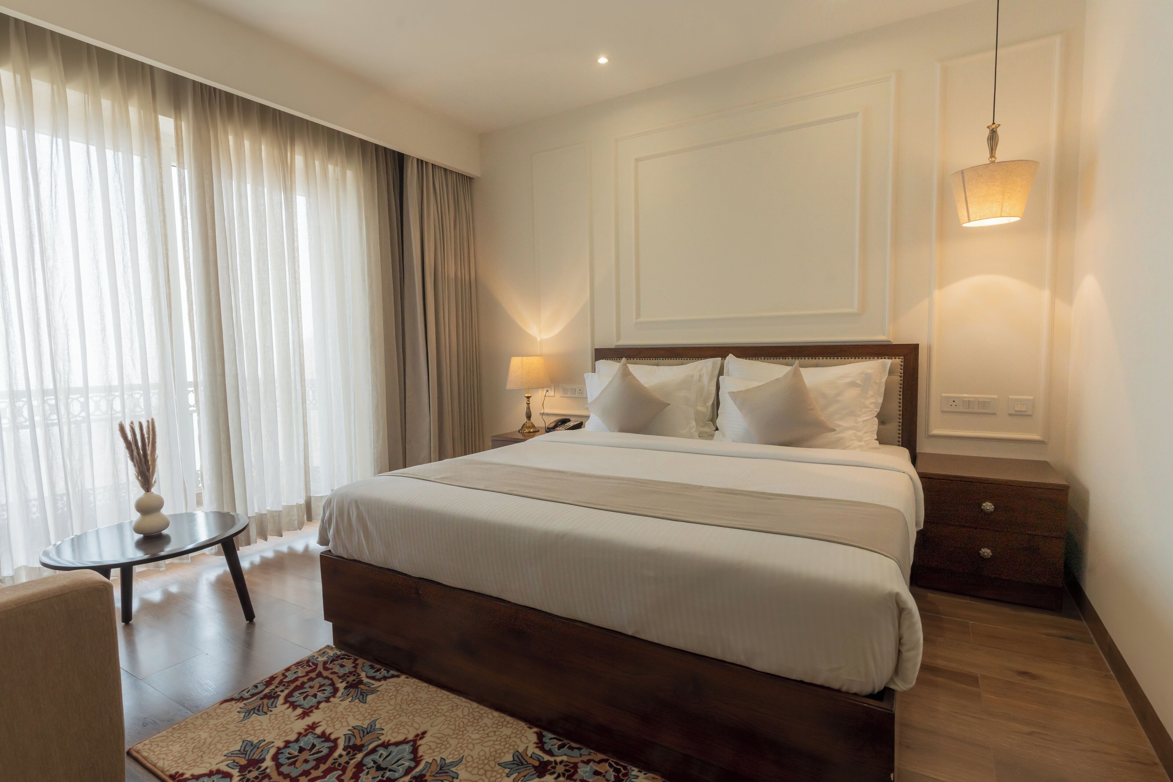A hotel room with a large bed, a bedside table with a lamp, a small round table with a vase, and a patterned rug.