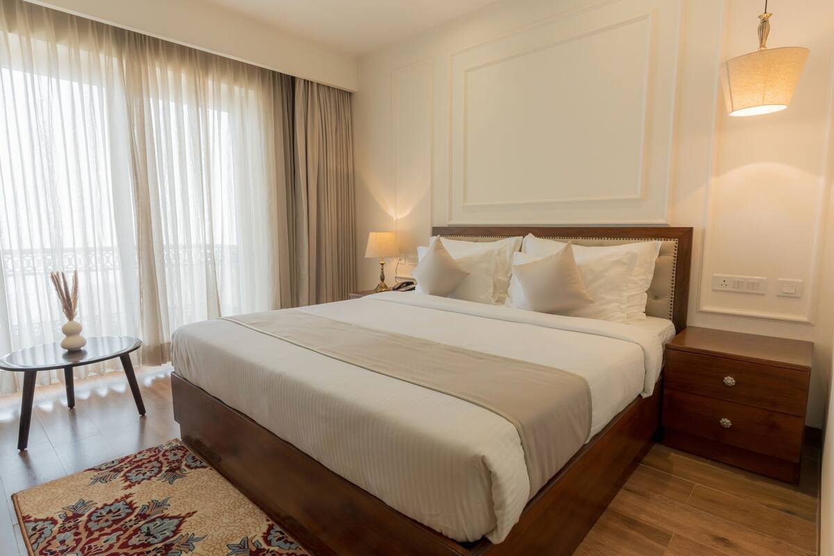 Hotelkamer met een groot bed, nachtkastje, lamp en uitzicht op het omliggende landschap door dunne gordijnen.