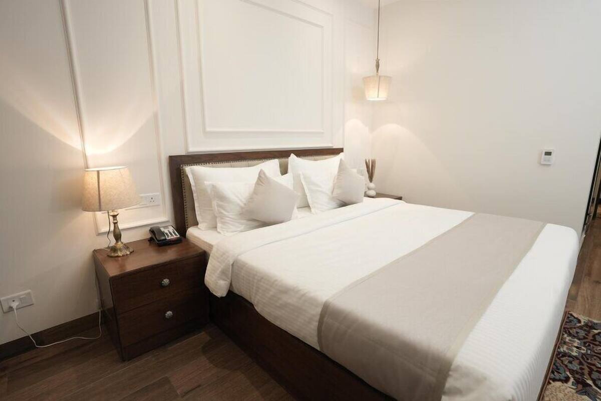 Een opgemaakt bed met witte lakens, een houten nachtkastje met een lamp en een wandlamp.