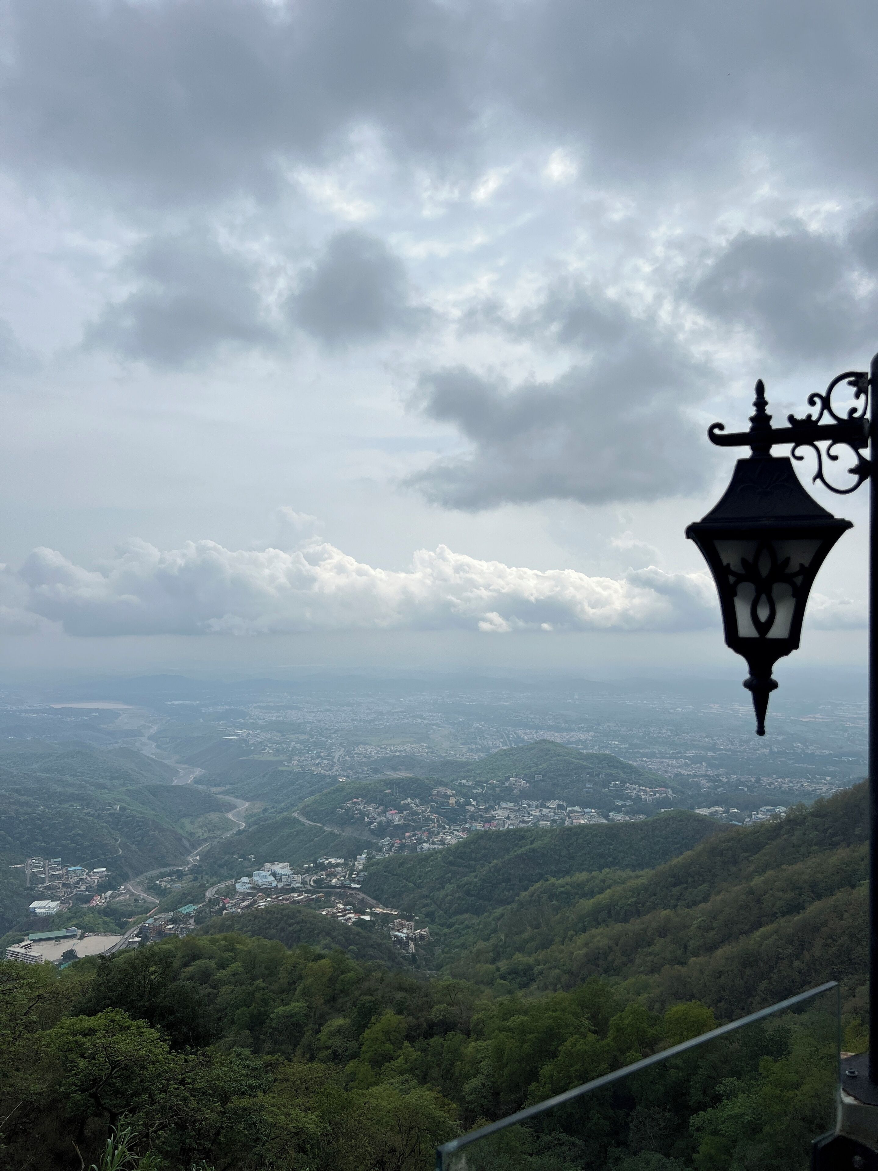 A view from a high vantage point, overlooking a valley with a town and greenery. A street lamp is visible in the foreground.