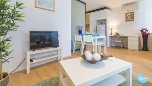 Un salon moderne avec un téléviseur à écran plat posé sur un meuble blanc, une table à manger avec des chaises, et un coin cuisine visible à travers une porte ouverte.