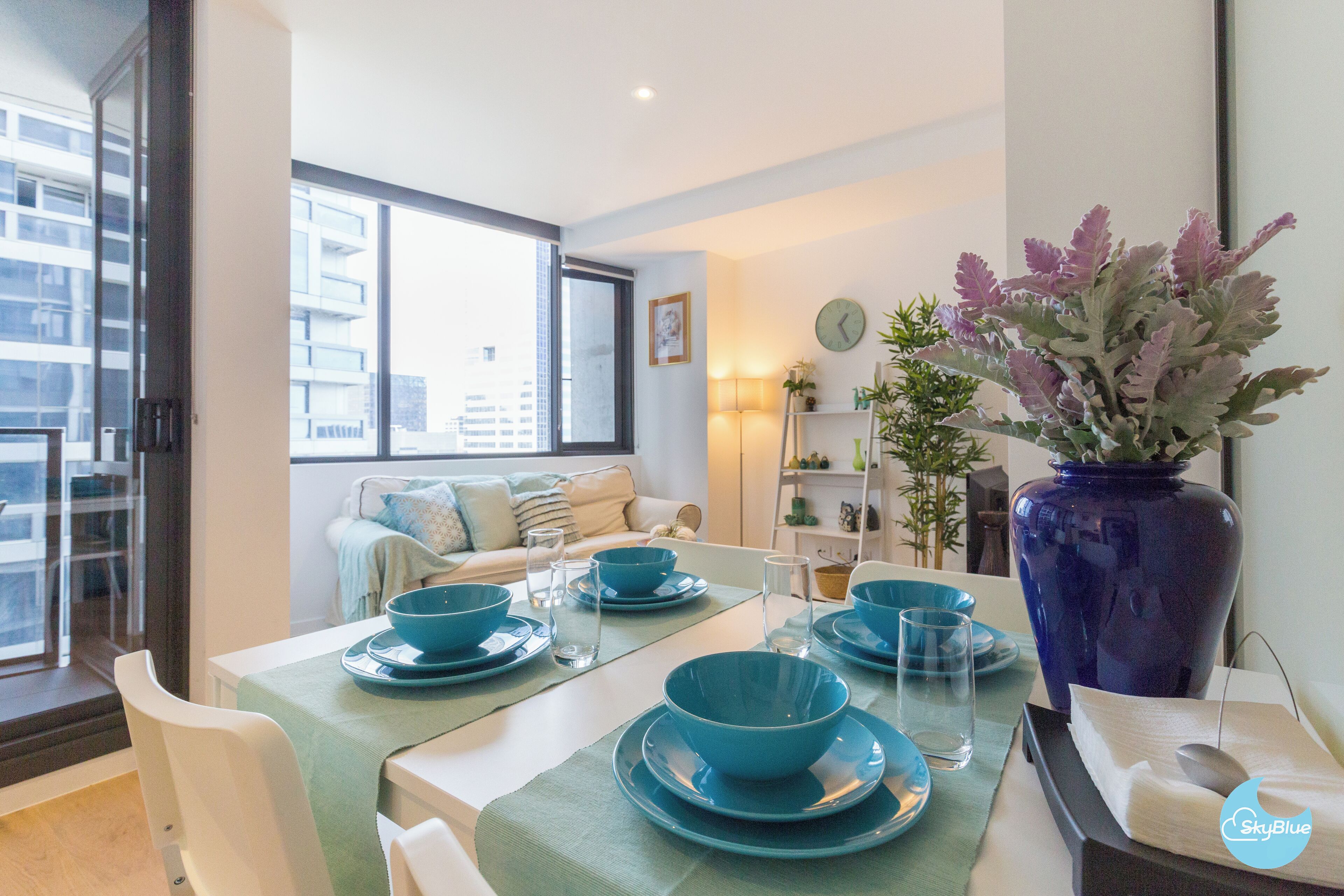 Uma sala de jantar com louças azul-turquesa, um vaso grande com flores e vista para a cidade através de uma janela ampla.
