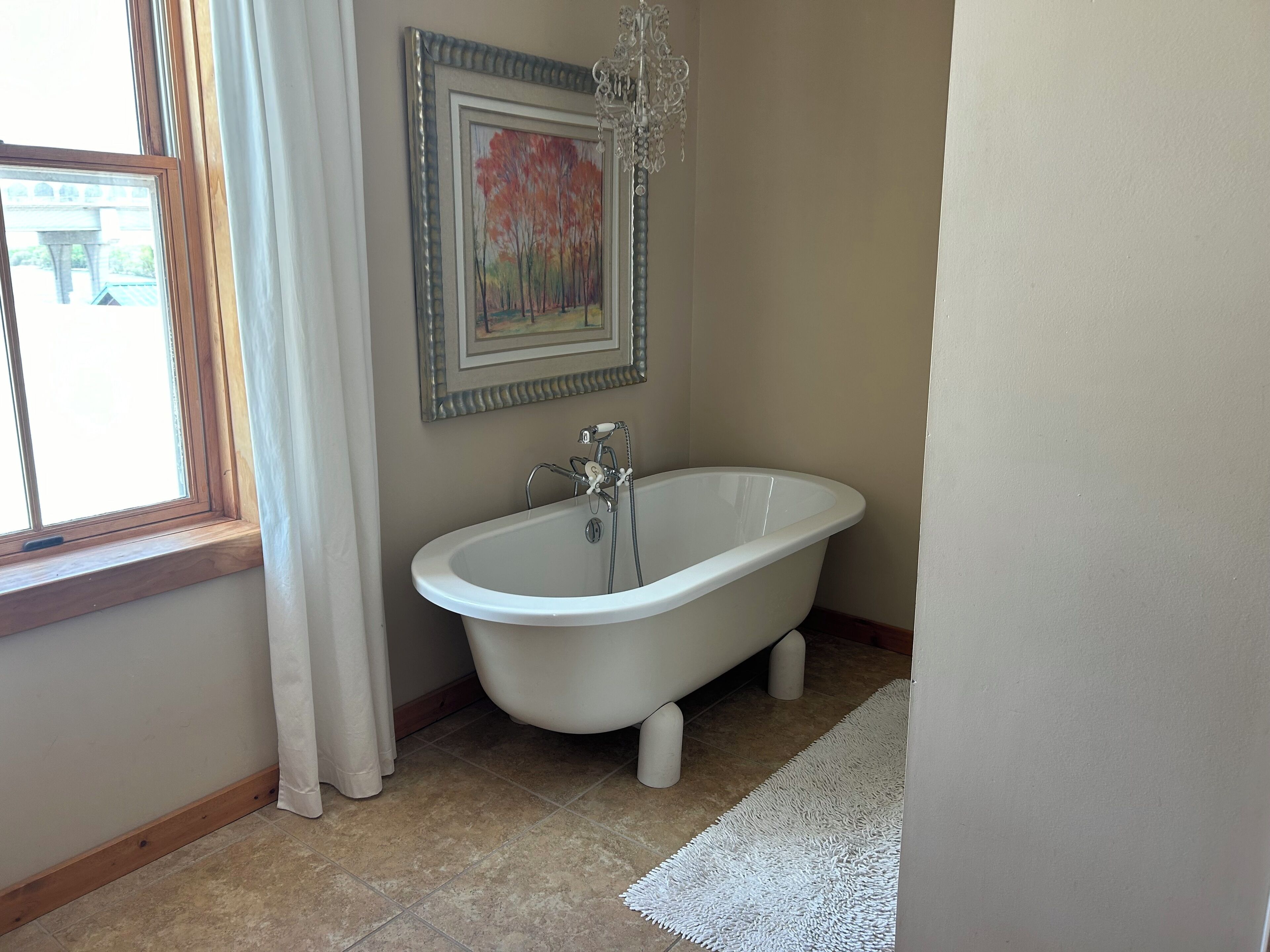 A bathroom with a freestanding bathtub, a window with curtains, a framed painting, and a chandelier.