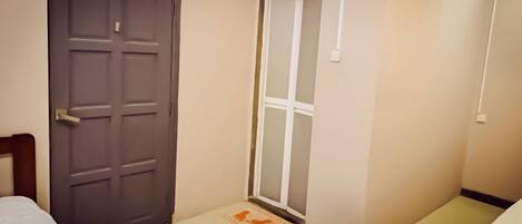 Um quarto pequeno e estreito, com uma porta, uma janela e uma cama.