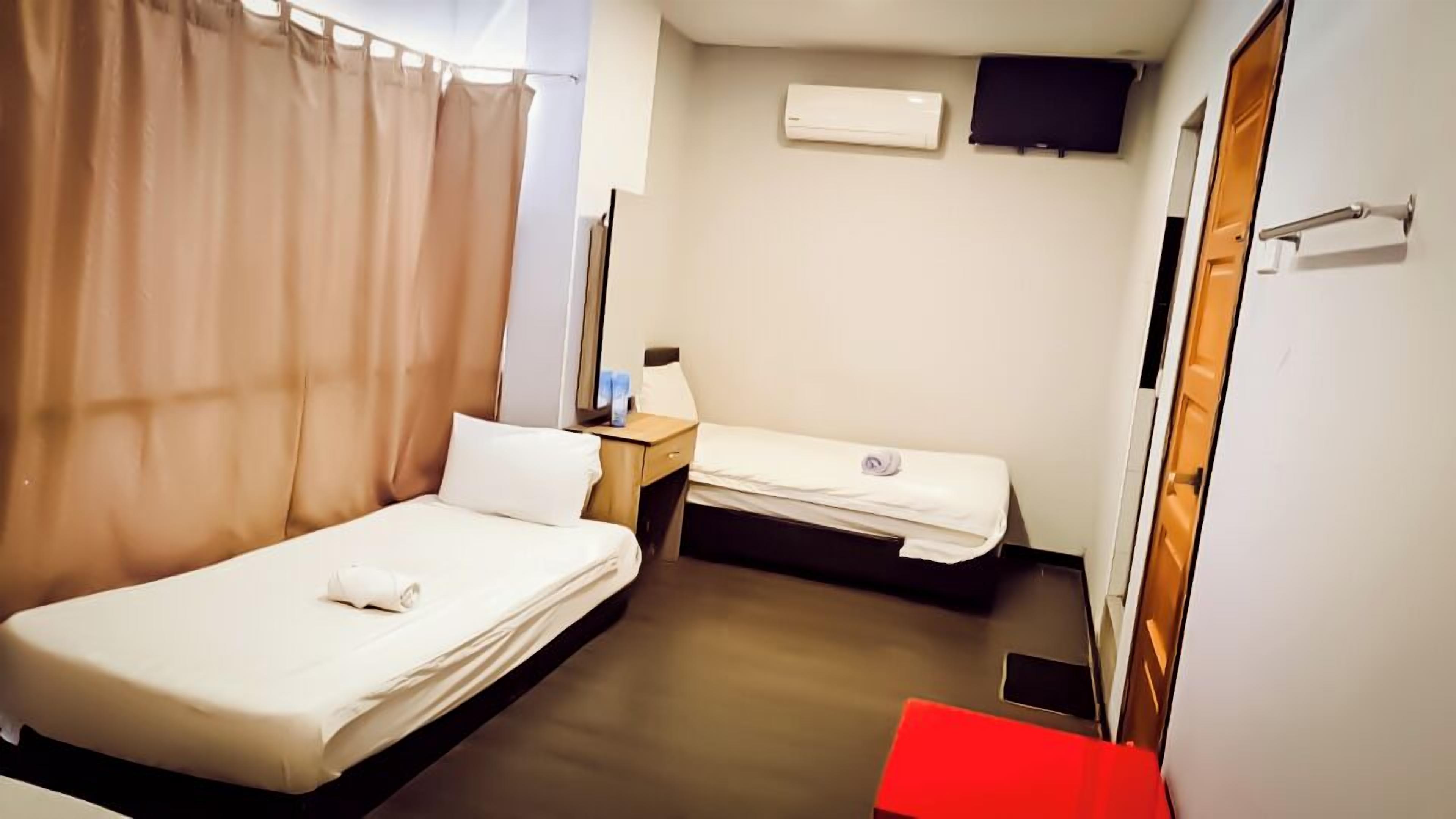 Ein kleines Hotelzimmer mit zwei Einzelbetten, einem Nachttisch mit Lampe, einem an der Wand montierten Fernseher und einer roten Bank.