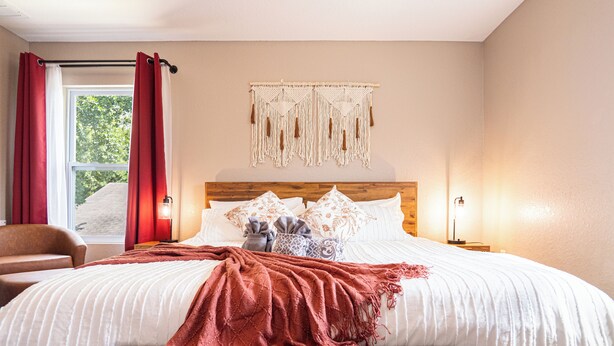 A bedroom with a large bed, a wooden headboard, a macrame wall hanging, and a view of trees through the window.