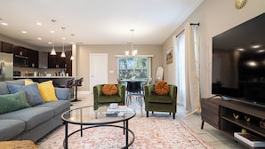 A modern living room with a sofa, two armchairs, a coffee table, a TV, and a kitchen in the background.