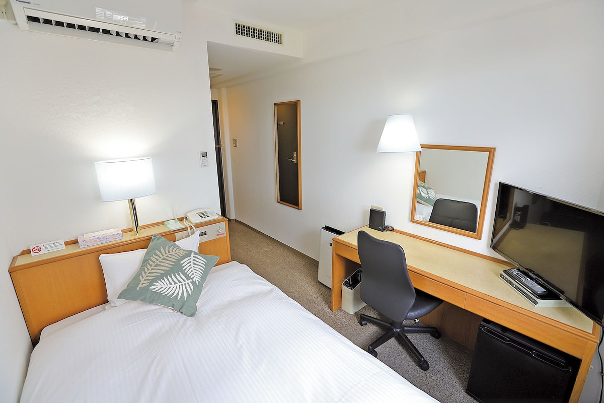 Habitación de hotel con cama, escritorio, silla, televisor, lámpara y espejo.