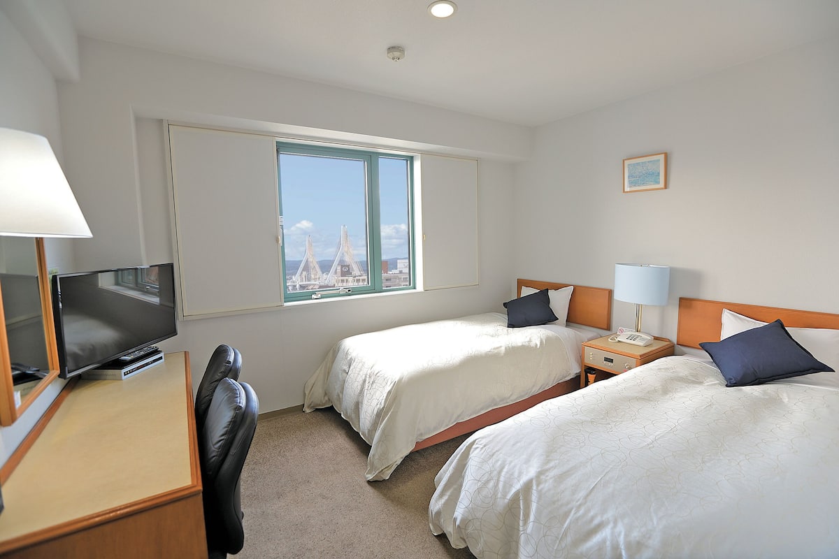 Habitación de hotel con dos camas, un escritorio con televisor, una silla y una ventana con vista a un puente.